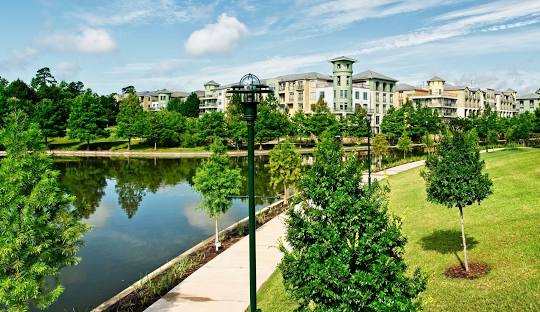 Lush green park with trees and a calm pond, featuring modern residential buildings in the background, emphasizing community and outdoor living in Harris County.