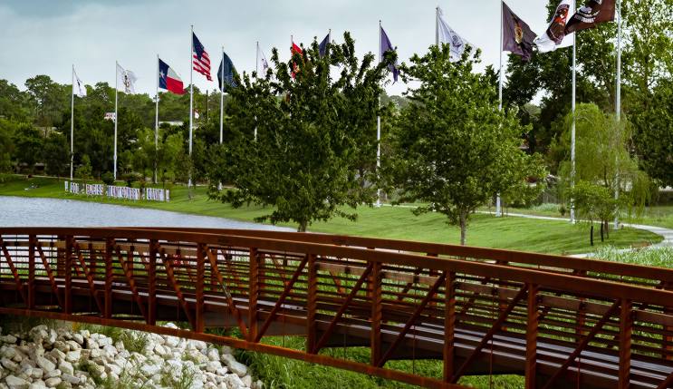 Bridge over a serene lake with trees and multiple flags representing different entities, symbolizing community and unity in Harris County, Texas.