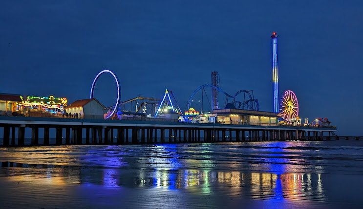 Colorful amusement park rides and attractions illuminated at night along a pier in Galveston, Texas, reflecting in the water.
