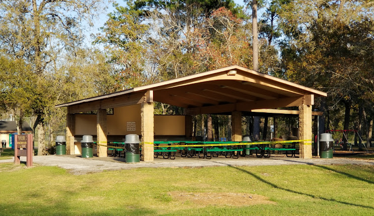 Pavilion with green and yellow caution tape, surrounded by trees, featuring picnic tables and trash bins, suitable for outdoor gatherings in a park setting.