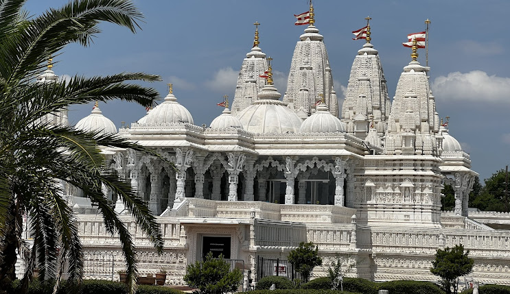 Ornate white temple with intricate carvings and multiple domes, surrounded by palm trees and landscaped gardens, under a bright blue sky.