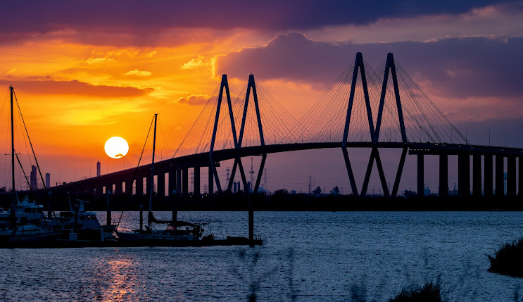 Sunset over a bridge with a vibrant orange and purple sky, boats in the foreground, symbolizing the scenic beauty of Harris County, Texas.