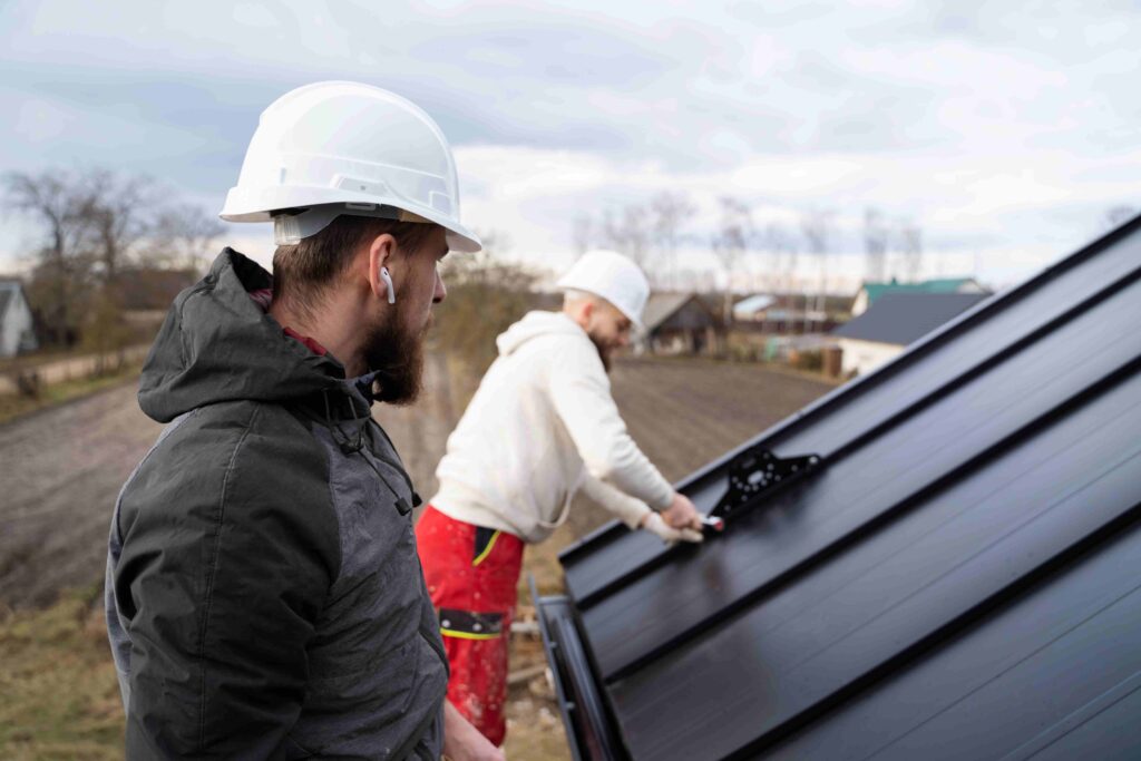 Two roofing professionals wearing safety helmets, one applying materials on a roof, while the other observes, set against a backdrop of a residential area, highlighting expert roof replacement services in Texas.