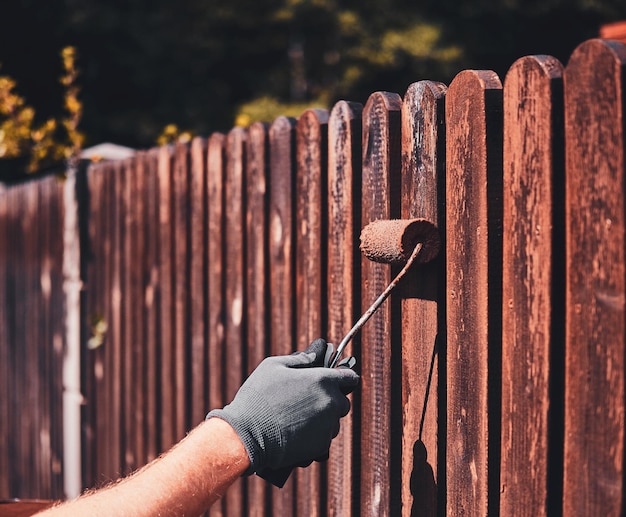 Hand applying protective stain to wooden fence with roller, emphasizing fence maintenance for durability in Texas weather.